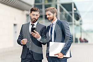 Two attractive smiling young businessmen wearing suits walking outdoors at the city streets, , using mobile phone