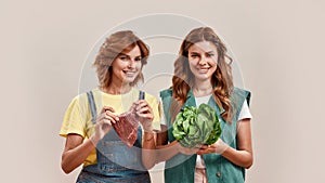 Two attractive young girls, twin sisters in casual wear holding fresh green salad and piece of meat isolated over light