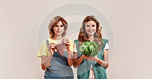 Two attractive young girls, twin sisters in casual wear holding fresh green salad and piece of meat isolated over light