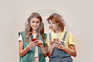 Two attractive young girls, twin sisters in casual wear using smartphone app while posing together, standing isolated