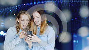 Two attractive young women enjoy a smartphone in a nightclub. They stand around of glass ornaments