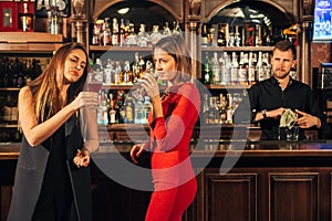 Two attractive young women meeting up in a pub for glass of red wine sitting at counter smiling each other