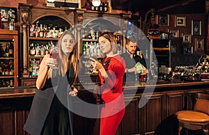 Two attractive young women meeting up in a pub for glass of red wine sitting at counter smiling each other