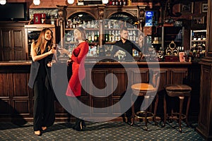 Two attractive young women meeting up in a pub for glass of red wine sitting at counter smiling each other