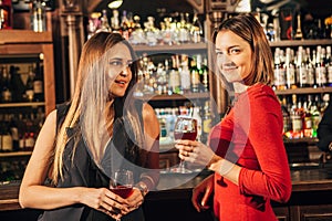 Two attractive young women meeting up in a pub for glass of red wine sitting at counter smiling each other