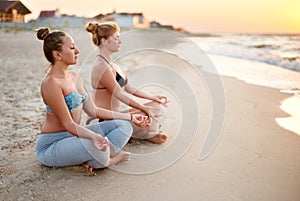 Two authentic women doing group yoga meditation on the beach at sunrise. Girls relaxing in lotus pose asana on sea shore