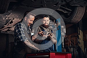 Two auto mechanic in a uniform, working on a workbench while standing under lifting car in the repair garage.