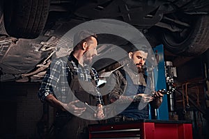 Two auto mechanic in a uniform, working on a workbench while standing under lifting car in the repair garage.