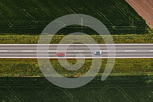 Two automobiles on straight asphalt highway through cultivated countryside landscape, aerial shot from drone pov