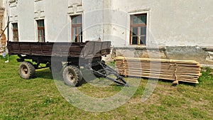 A two-axle metal trailer with sides and drawbar stands partially on the grassy lawn near the wall of a building with wooden window