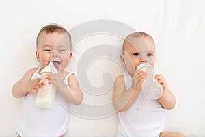 Two babies suck milk from a bottle lying in a crib in the children`s room on a white bed
