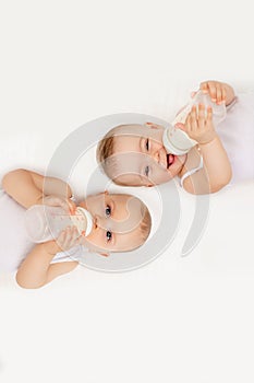 Two baby twins boy and girl with a bottle of milk on a white bed at home, baby food concept, place for text