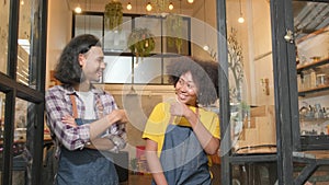 Two baristas at a cafe door, arms crossed, teasing together with a happy smile.