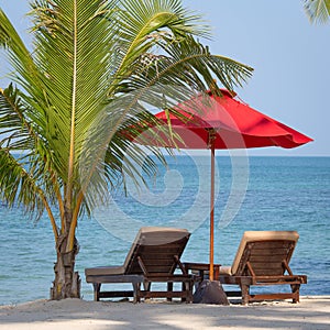 Two beach chairs, red umbrella and palm tree on the beach in Thailand