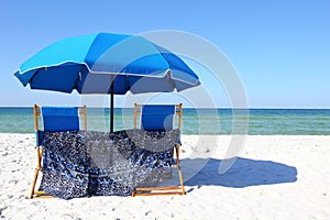 Two beach chairs under a blue umbrella on a white sandy beach