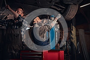 Two bearded auto mechanic in a uniform repair the car`s suspension while standing under lifting car in the repair garage