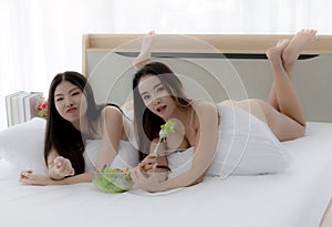 Two beautiful Asian women in a sheer costume lie on a white couch eating healthy food. A sexy young woman holding a small party