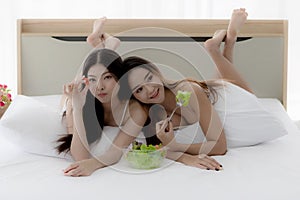 Two beautiful Asian women in a sheer costume lie on a white couch eating healthy food. A sexy young woman holding a small party