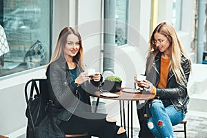 Two beautiful attractive stylish women are sitting outdoor in cafe drinking coffe and tea talking and enjoying great day.
