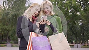 Two beautiful blond girlfriends standing on the street with shopping bags looking at cellphone. Two fashion women