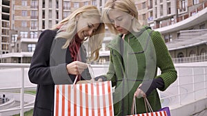 Two beautiful blond girlfriends standing on the street showing each other shopping bags. Two fashion women show off