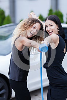 Two beautiful businesswomen in black outfut posing in front of an electric car. Girls holding blue charging cable. Shallow depth