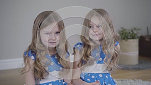 Two beautiful Caucasian twin girls sitting at home and holding hands. Portrait of little brunette sisters indoors. Unity