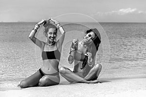 Two beautiful cheerful young women twin sisters in bikini sitting on sand on the beach at sunny day