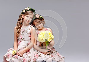 Two beautiful cute girls - sisters in beautiful dresses in wreaths of fresh flowers on their heads are sitting and hugging in the