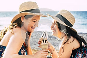 Two beautiful female friends in straw hat having fun enjoying cold ice cream cones on a peaceful beach during summer holiday.