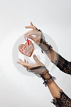 Two beautiful female hands in black lace gloves hold a heart shaped gingerbread cookie on red ribbon