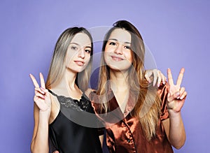 two beautiful females dressed in pajamas hugging and show peace sign over purple background.