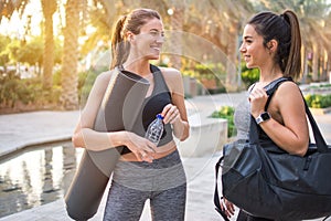 Two beautiful fitness female friends in sportswear holding gym bag, yoga mat and water bottle talking in the park.