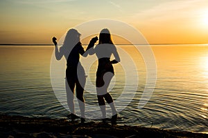 Two beautiful girls are dancing holding hands on the beach at sunset background, silhouettes