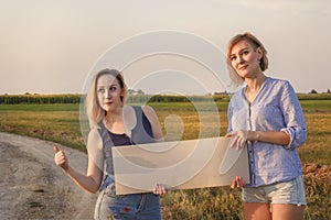 Two beautiful girls get hitchhiked and vote with a sign on cars on a rural road in the rays of sunset.