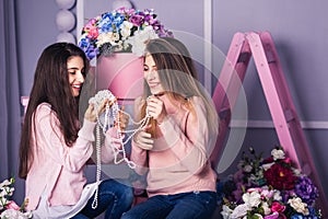 Two beautiful girls in jeans and pink sweaters are holding beads in studio with decor of flowers in baskets.