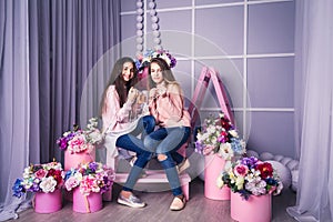 Two beautiful girls in jeans and pink sweaters are holding beads in studio with decor of flowers in baskets.