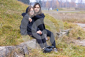 Two beautiful girls with long hair in black clothes sit together on the grass on the outskirts of the city