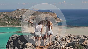 Two beautiful girls standing on edge of cliff. Female friends admire view of blue sea laggon and rocky beach, pointing