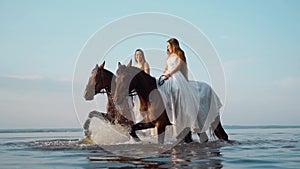 Two beautiful girls in white long dresses on horseback. Horse riders stroll through the water along the coast at sunset.