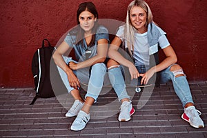 Two beautiful hipster girls sitting on a skateboard and leaning on a wall.