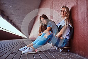 Two beautiful hipster girls sitting on skateboard at a sidewalk under bridge.