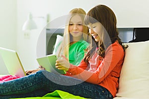 Two beautiful little sisters sitting on bed and play with a Tablet or laptop