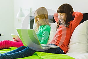 Two beautiful little sisters sitting on bed and play with a Tablet or laptop