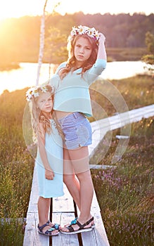 Two beautiful sisters standing against the background of a beautiful landscape, walk on the field near a pond at sunset.