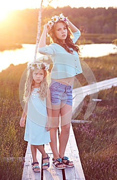 Two beautiful sisters standing against the background of a beautiful landscape, walk on the field near a pond at sunset.