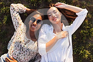 Two beautiful smiling girls lying on grass while happily spending time together in city park