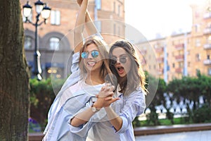 Two beautiful smiling girls in trendy summer clothes posing on street background. Models are having fun and hugging