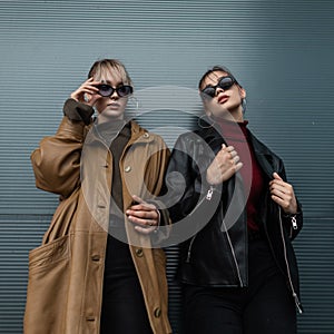 Two beautiful teenage model women in a leather black jacket with black jeans wearing sunglasses poses near a metal wall