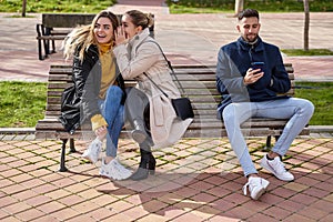 Two beautiful teenagers share secrets, gossip while a boy looks at his cell phone, they are sitting on a park bench